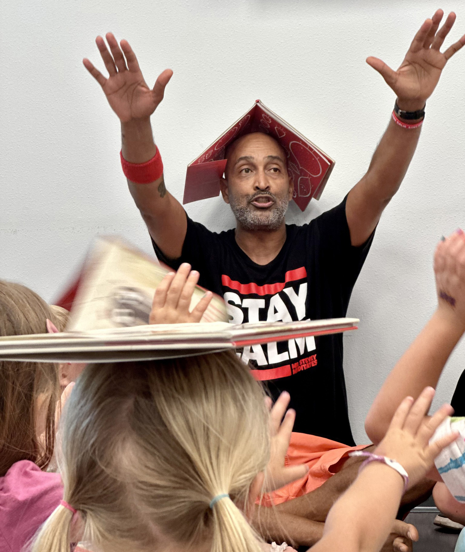 Mr. Stevey leads children in an interactive storytime activity, raising his hands while wearing a book on his head as children participate around him.