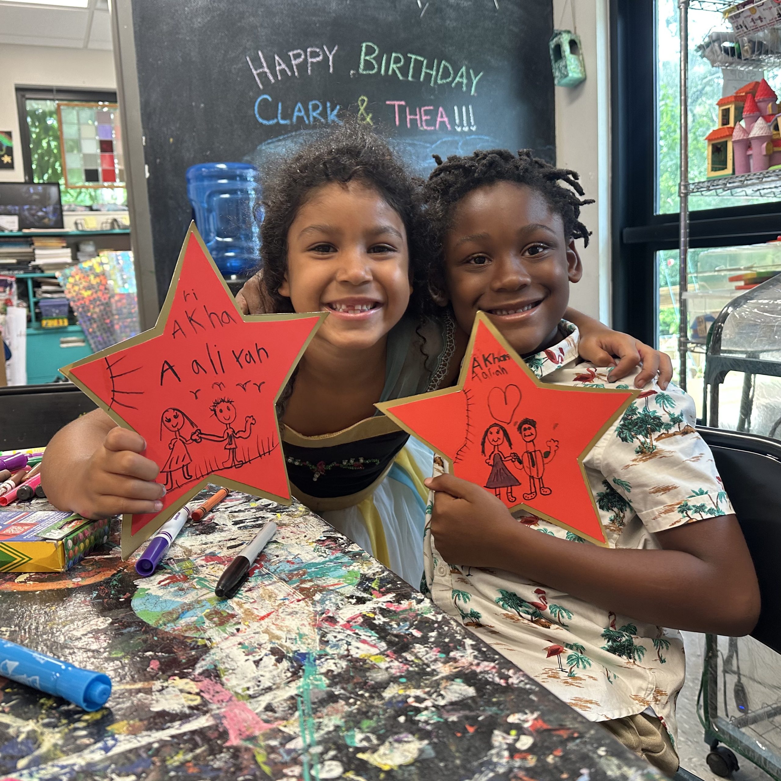 Two smiling children sit together and display handmade red stars during a Community Stepping Stones art activity.